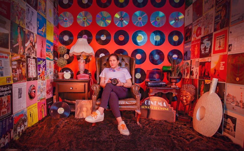 Seated on a velvet sofa against a red wall of vinyl records and a glowing cinema sign