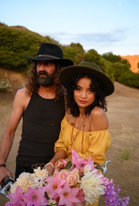 Two people in wide-brim hats with a bouquet of flowers, photographed in golden light
