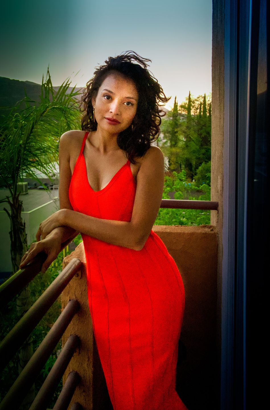 Woman in a red dress leaning on a balcony rail surrounded by greenery