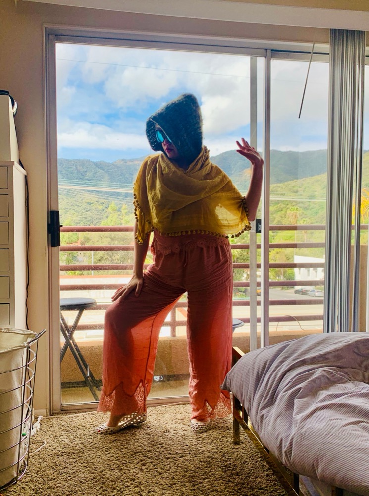 Figure in a yellow shawl and headwrap striking a playful pose by a sunlit window